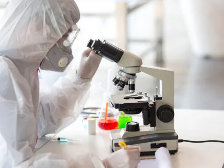 Wissenschaftler im Sicherheitsanzug, der biologische Flüssigkeiten unter dem Mikroskop betrachtet